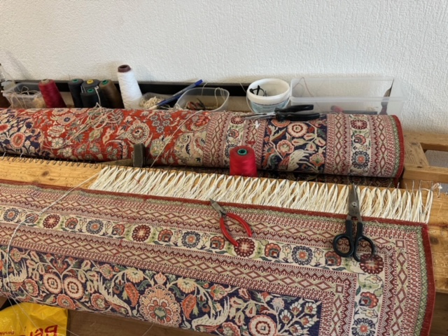 Frange neuve en fils blancs tendue autour des chevilles d'un métier de bois, un tapis persan rouge en cours de réfection et ses outils posés dessus