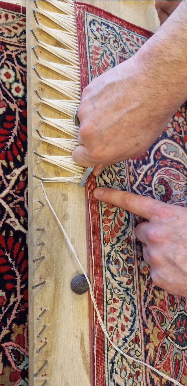 Les mains d'un artisan resserrent à l'outil la base d'une frange neuve en fils blancs, le long de la bordure rouge d'un tapis persan