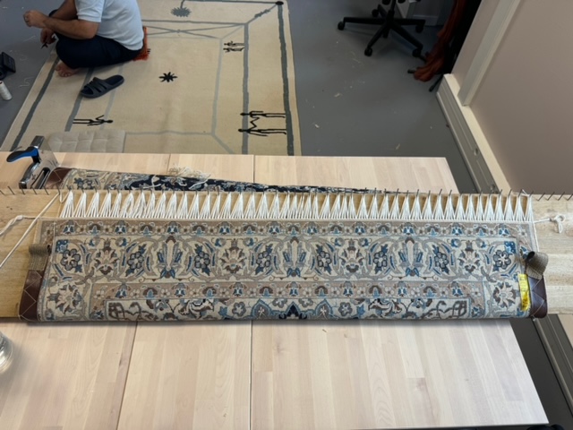 Dans l'atelier, un grand tapis roulé dont la frange neuve est tendue en réguliers brins blancs autour des chevilles du cadre de bois