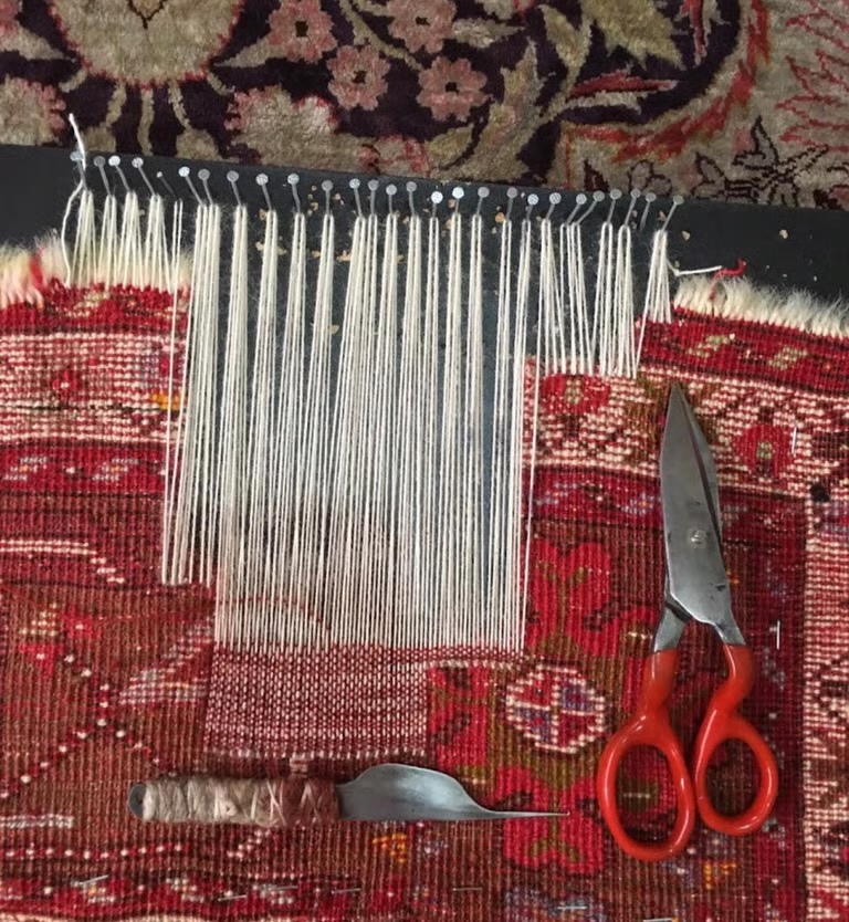 Chaîne reconstituée brin par brin et tendue sur des épingles, avec les ciseaux et l'outil de l'artisan posés sur le tapis